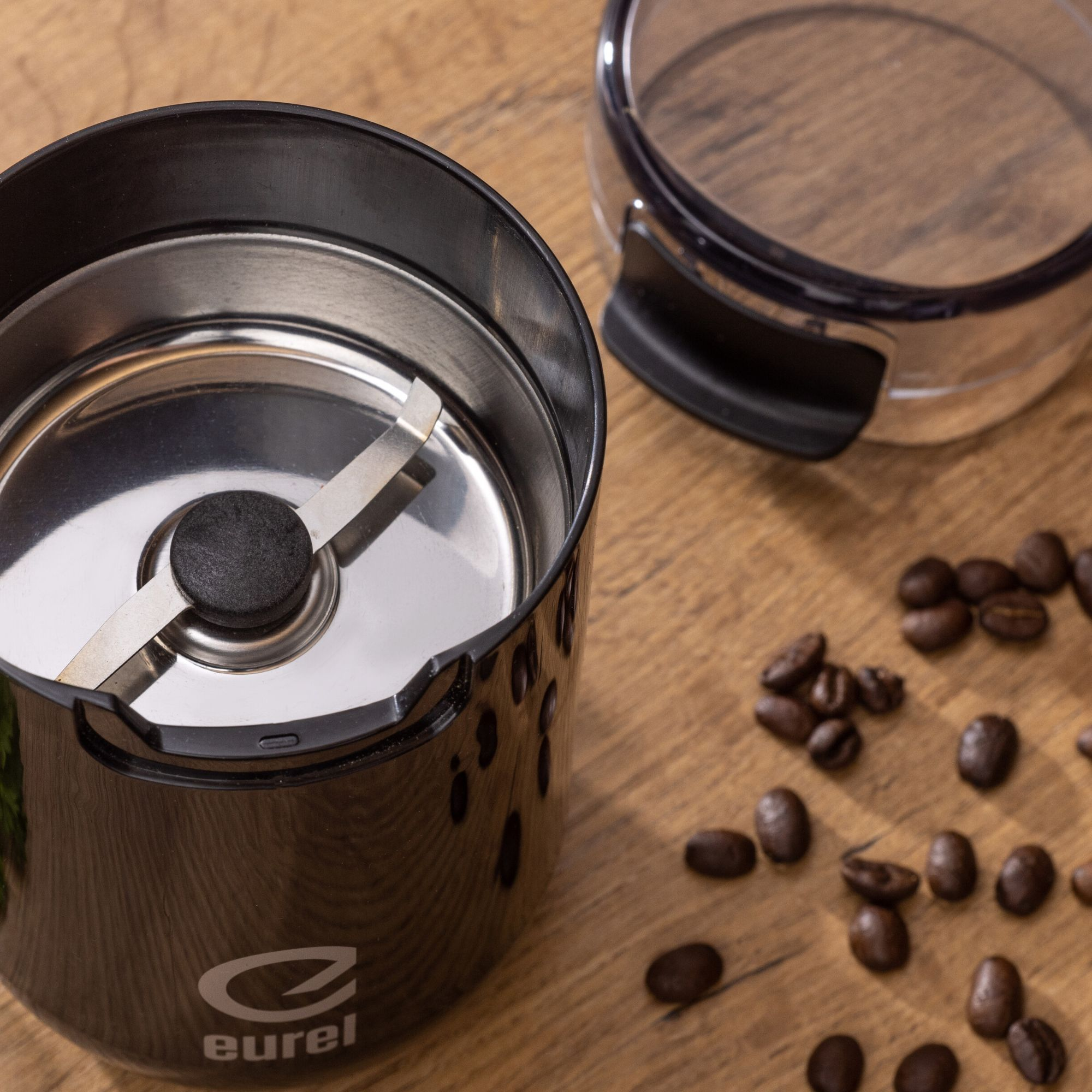 Schwarze Kaffeemühle mit Kaffeebohnen auf einem Holztisch verstreut, klarer Deckel oben.