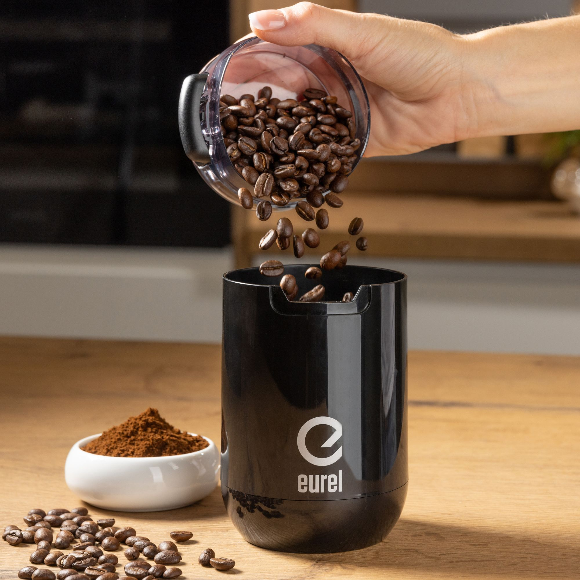 Eine Hand gießt Kaffeebohnen in eine schwarze elektrische Kaffeemühle, mit Kaffeepulver.