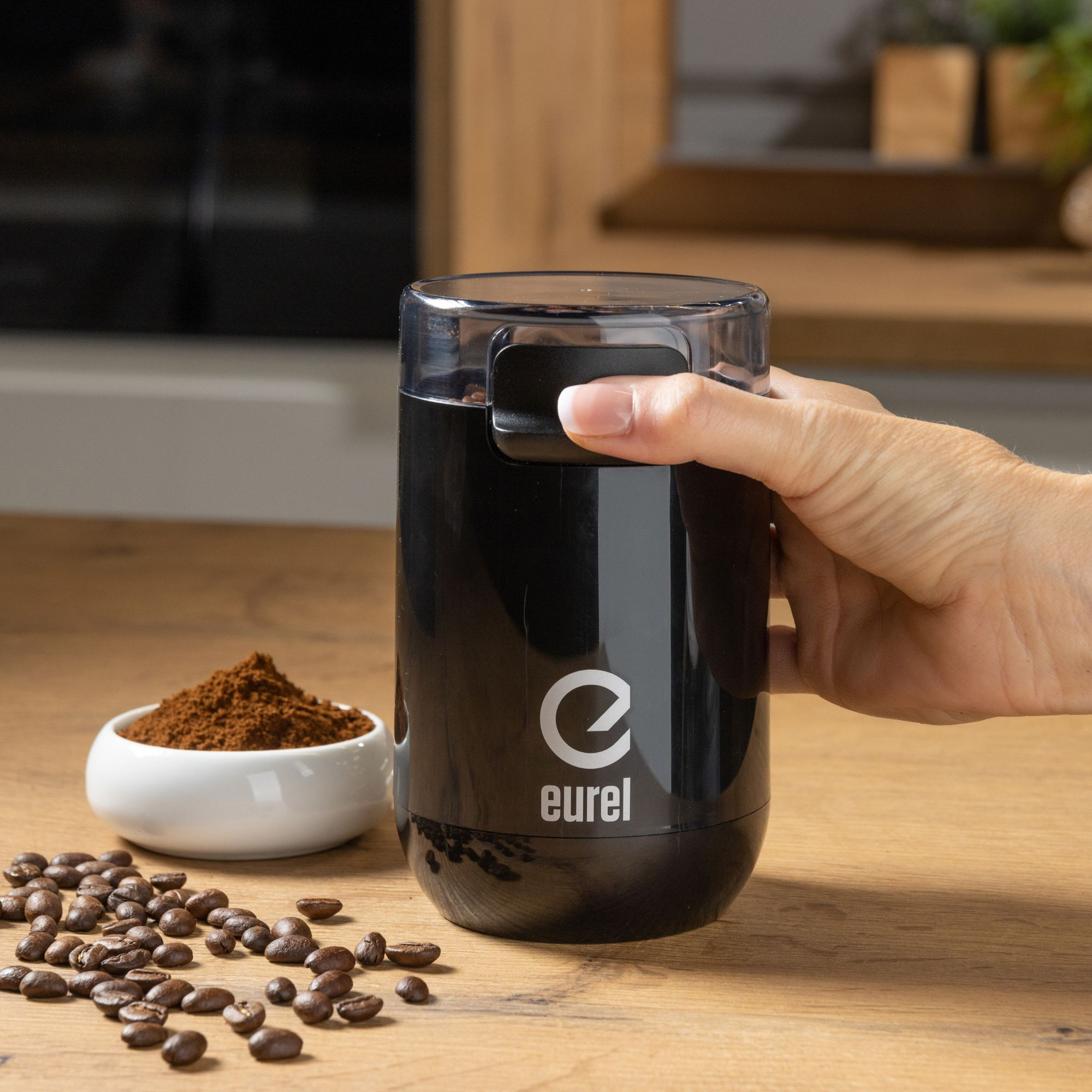 Eine schwarze Kaffeemühle mit einer Hand obenauf, neben einer Schüssel mit Kaffeepulver und Kaffeebohnen.