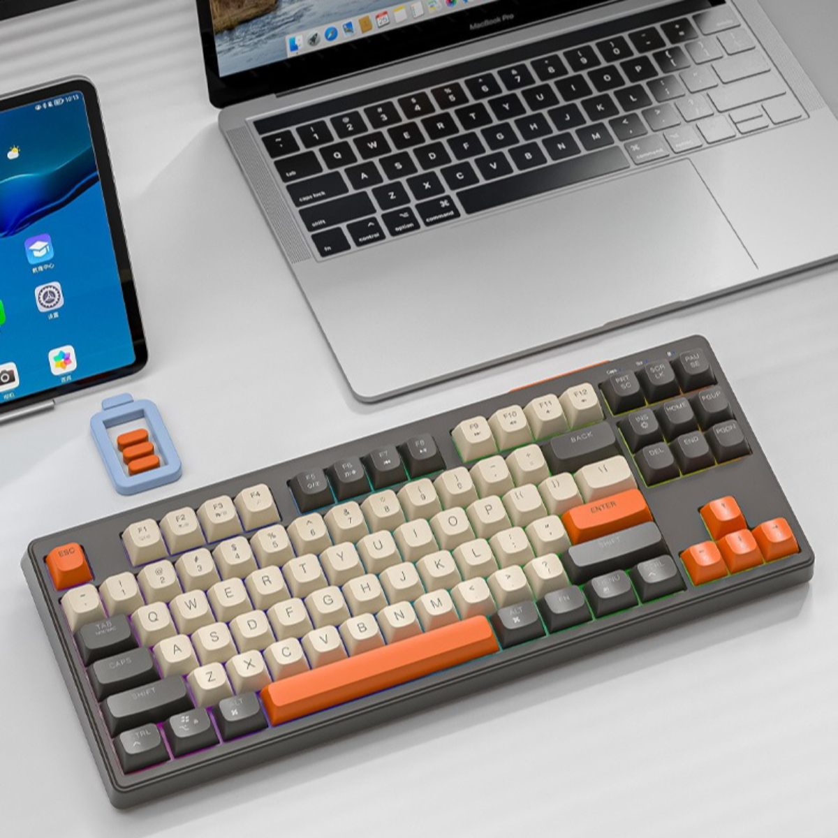 Eine mechanische Tastatur mit orangefarbenen und grauen Tasten, ein Tablet und ein Laptop auf einem weißen Tisch.