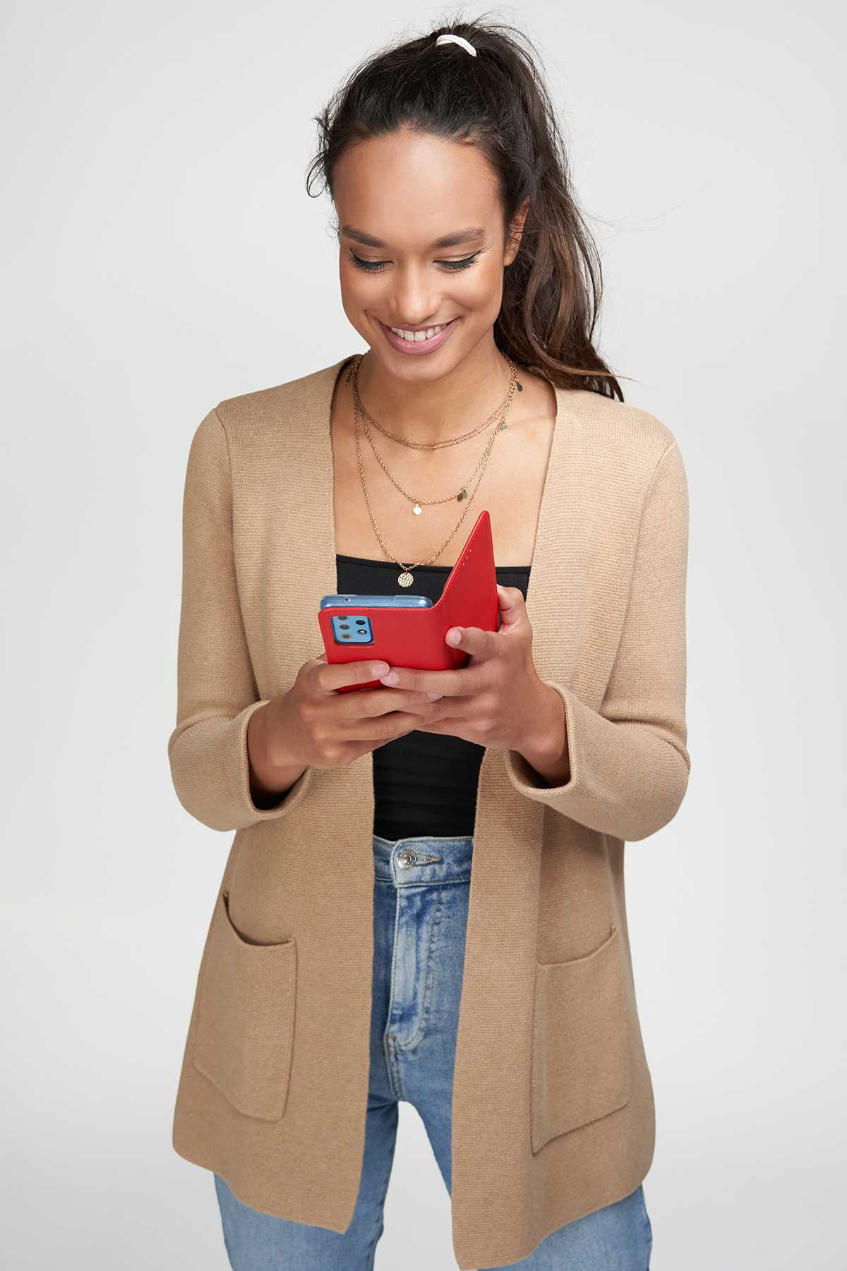 Frau in beigefarbener Strickjacke hält rotes Telefon, lächelt. Jeans, schwarzes Top. Hintergrund ist schlicht.
