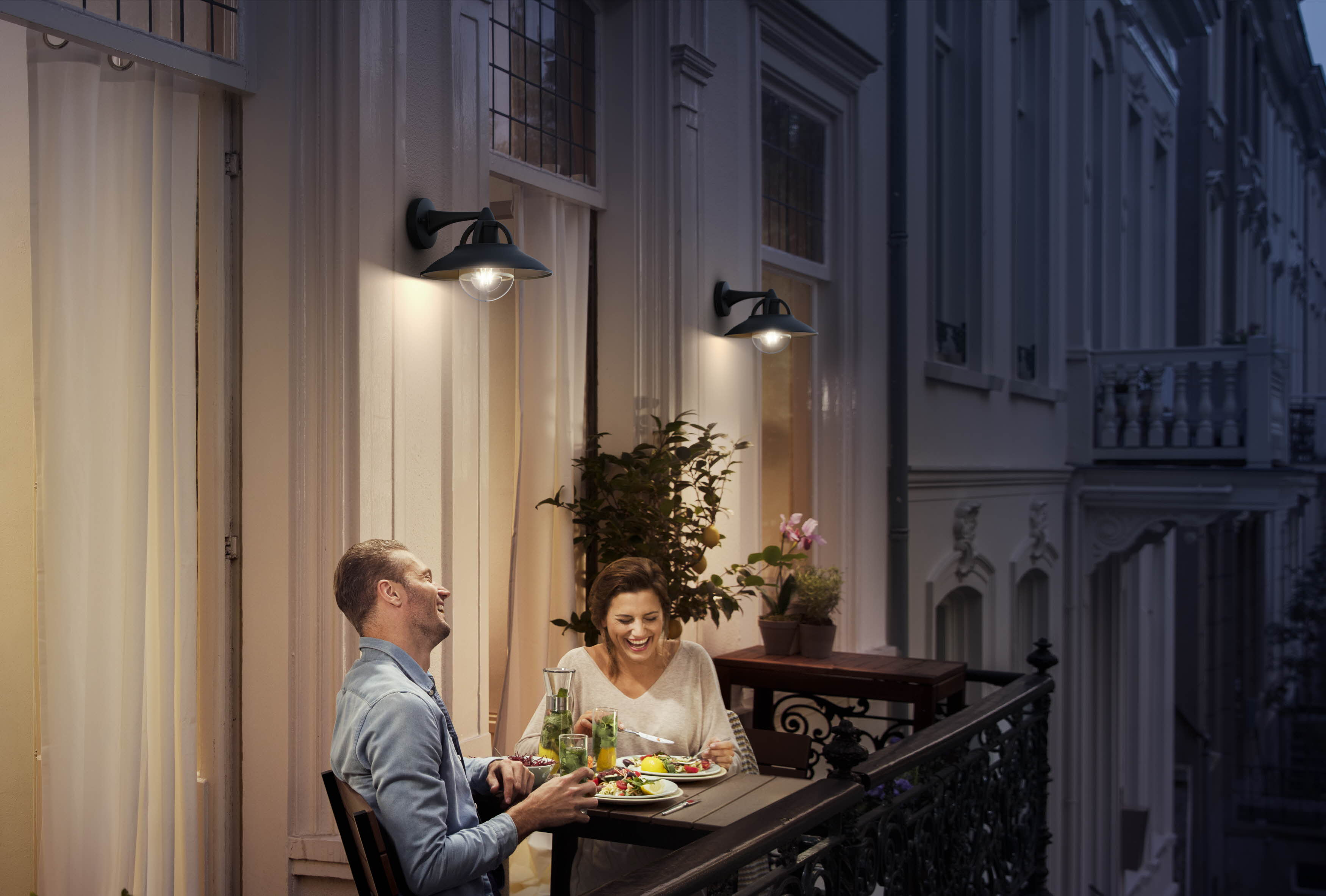 Paar auf einem Balkon bei Nacht. Sie sitzen an einem Tisch und essen. Zwei Lampen an der Wand.