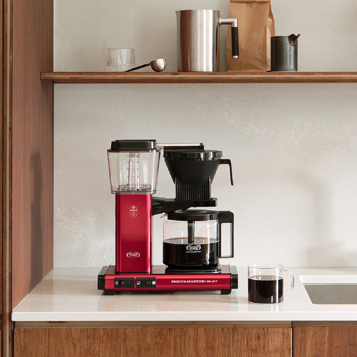 Cafetera roja sobre una encimera con café e ingredientes en una estantería.