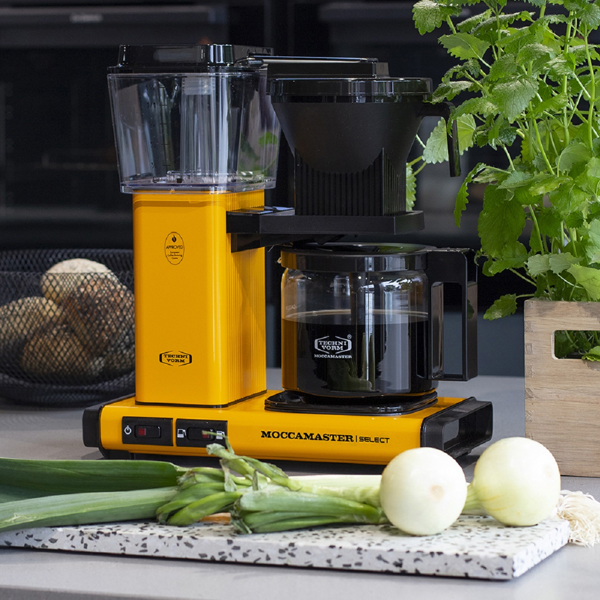 Yellow Moccamaster coffee maker with clear water reservoir, black filter basket, and glass carafe on countertop.