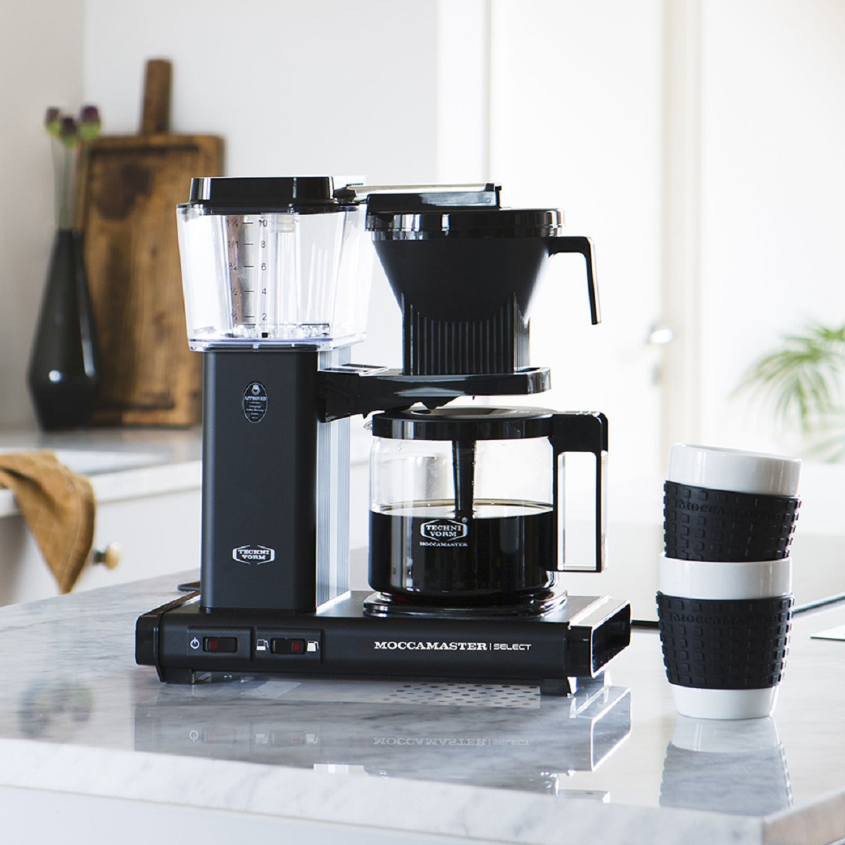 Zwarte Moccamaster koffiezetapparaat met helder waterreservoir, zwart filtermandje, glazen kan. Twee mokken rechts.