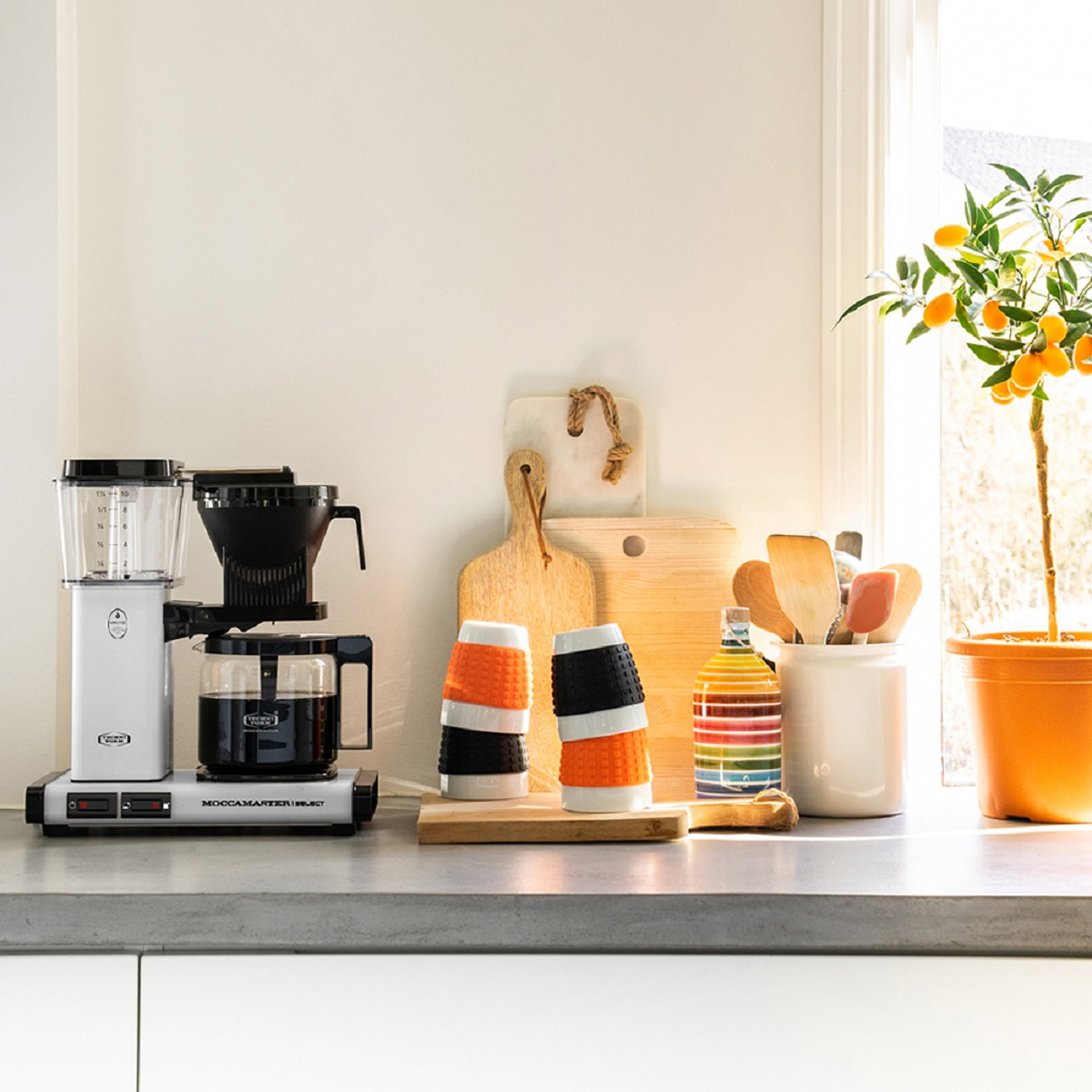 Scena kuchenna z ekspresem do kawy, deskami do krojenia, kubkami i małą rośliną.