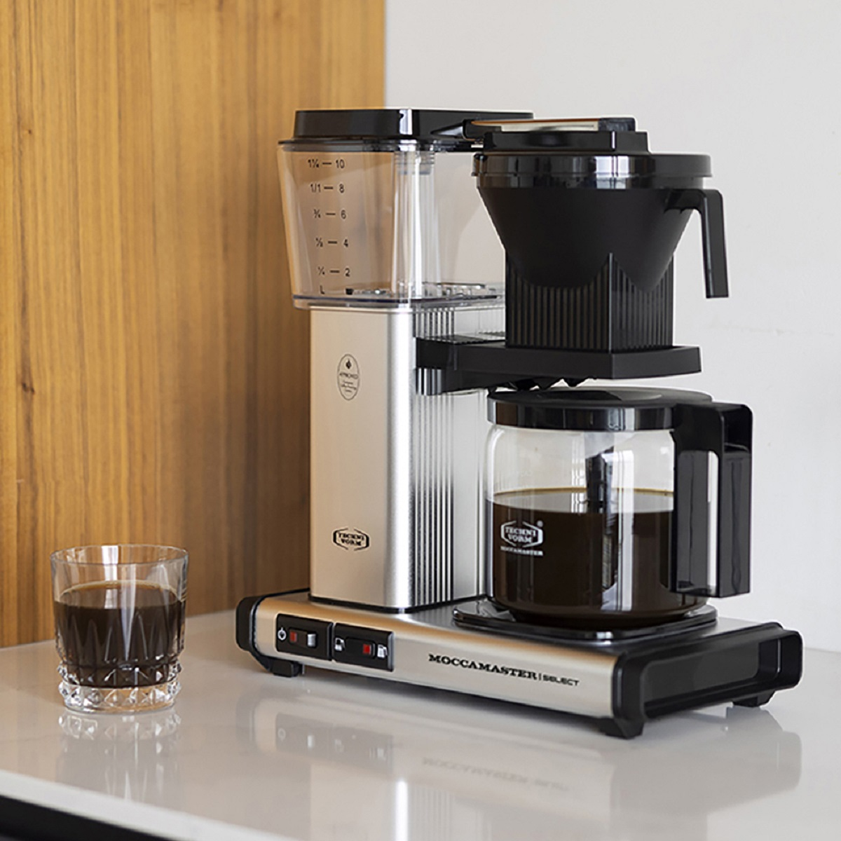 Coffee machine with a glass carafe, a glass of coffee, and a wooden panel background.