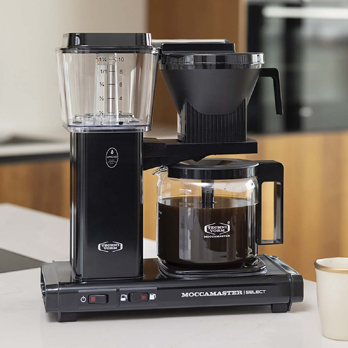 Czarny ekspres do kawy ze szklaną karafką i przezroczystym pojemnikiem na wodę. Stoi na blacie.