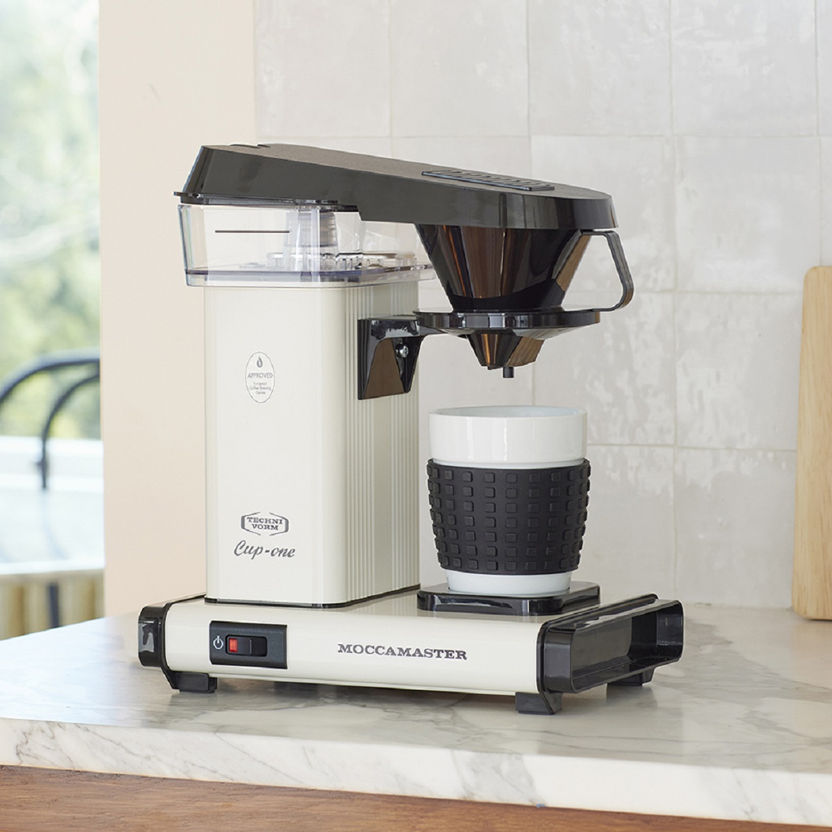 Eine cremefarbene Kaffeemaschine mit schwarzem Griff und einer weißen Tasse auf der Arbeitsplatte.