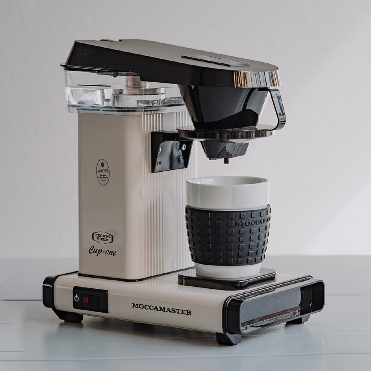Creme-schwarze Kaffeemaschine mit einer Tasse. Der Text lautet: Cup-one und Moccamaster.