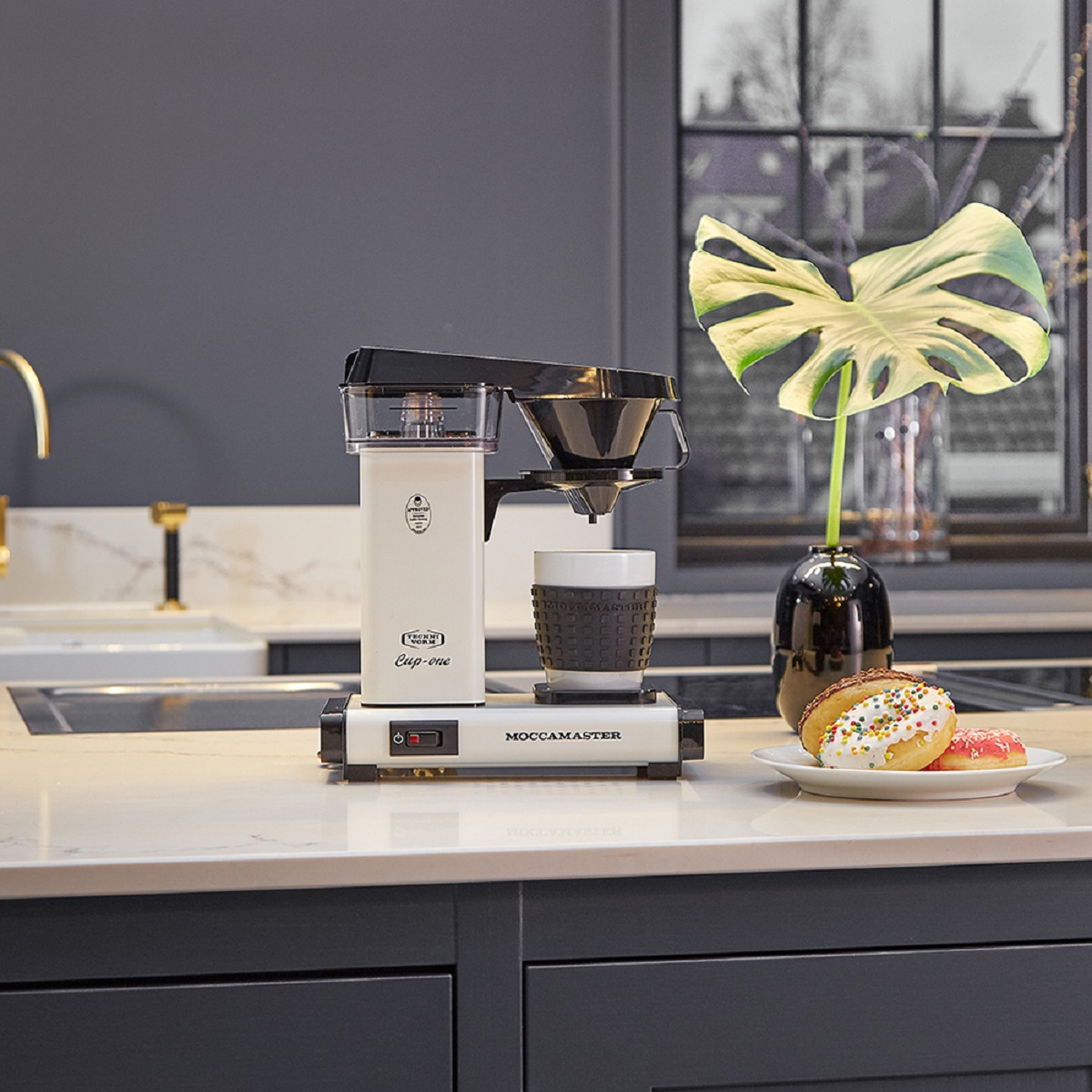 A white coffee maker with a cup, donuts, and a plant on a countertop.