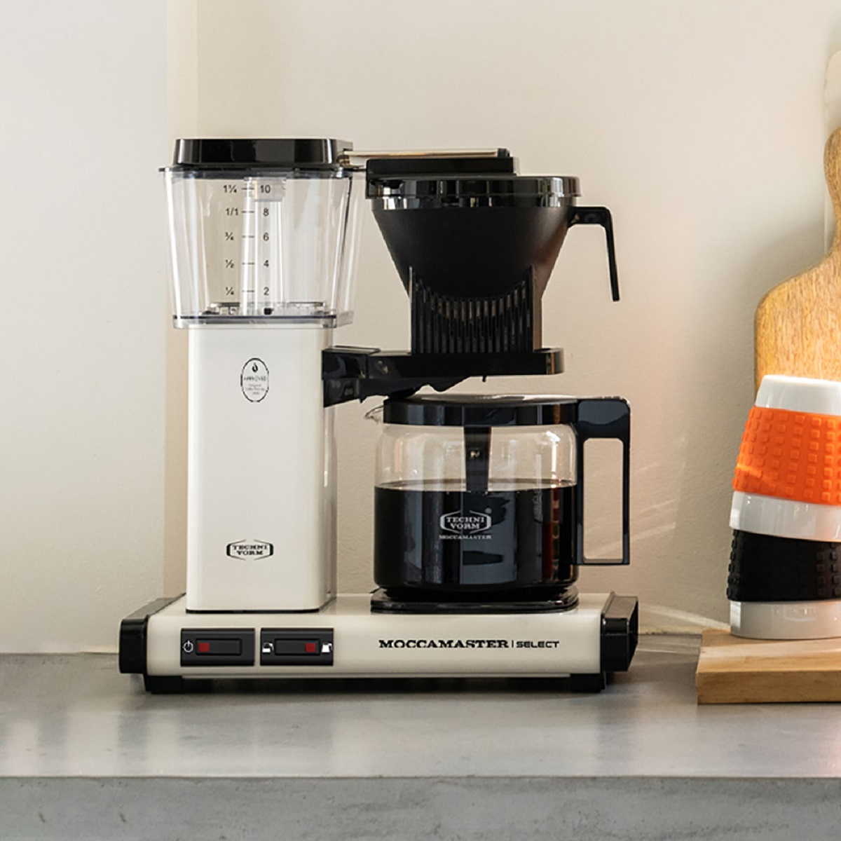 A white coffee maker. It has a glass carafe and a black filter holder. The background is a kitchen counter.