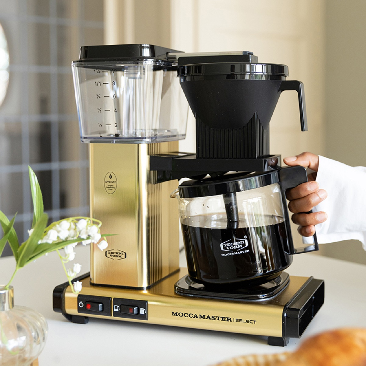 Eine gold-schwarze Kaffeemaschine mit einer Glaskaraffe gefüllt mit Kaffee. Eine Hand gießt Kaffee ein.