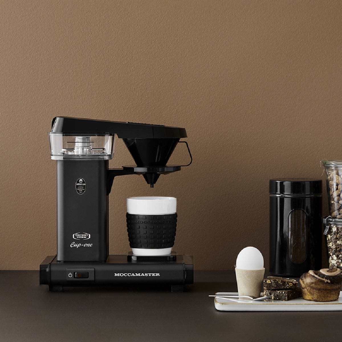 Black coffee maker with a cup and pastries on a table with various ingredients.