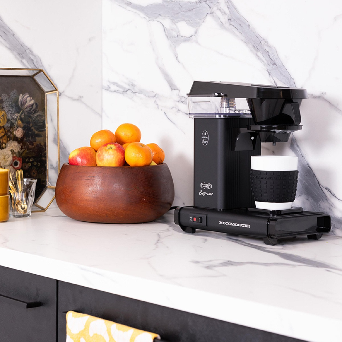 A black coffee maker with a cup, and a bowl of oranges on a white countertop.