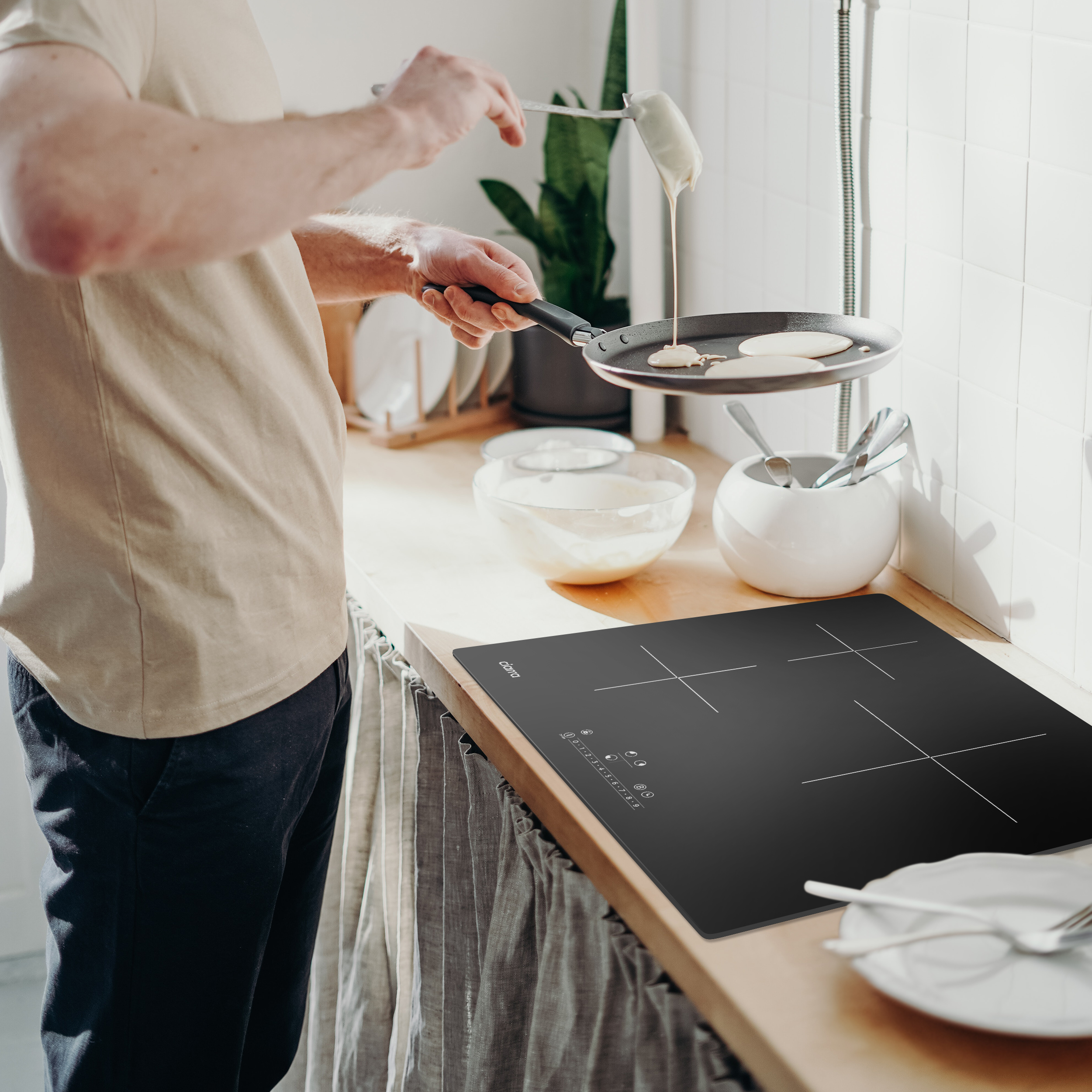 Une personne faisant des crêpes sur une plaque de cuisson noire. Scène de cuisine avec ustensiles et ingrédients.