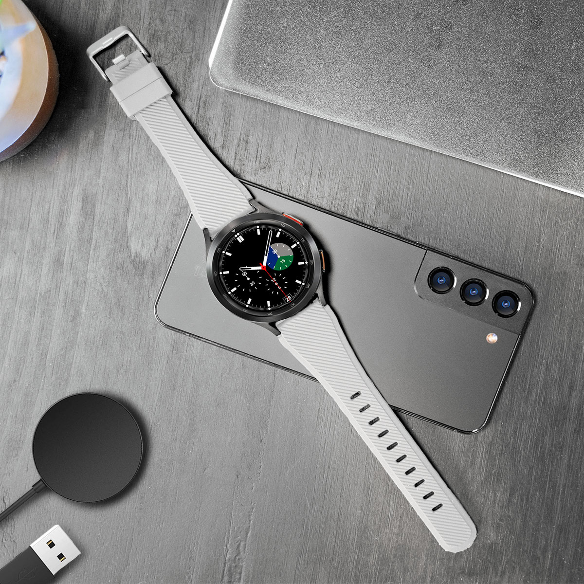 Smartwatch mit grauem Armband, Smartphone, Ladegerät und USB-Kabel auf Holzoberfläche.
