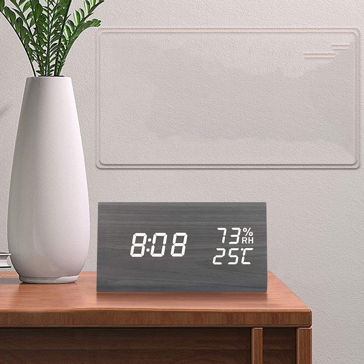 Holzuhr mit Digitalanzeige, die Zeit, Temperatur und Luftfeuchtigkeit anzeigt. Weiße Vase und Pflanze im Hintergrund.