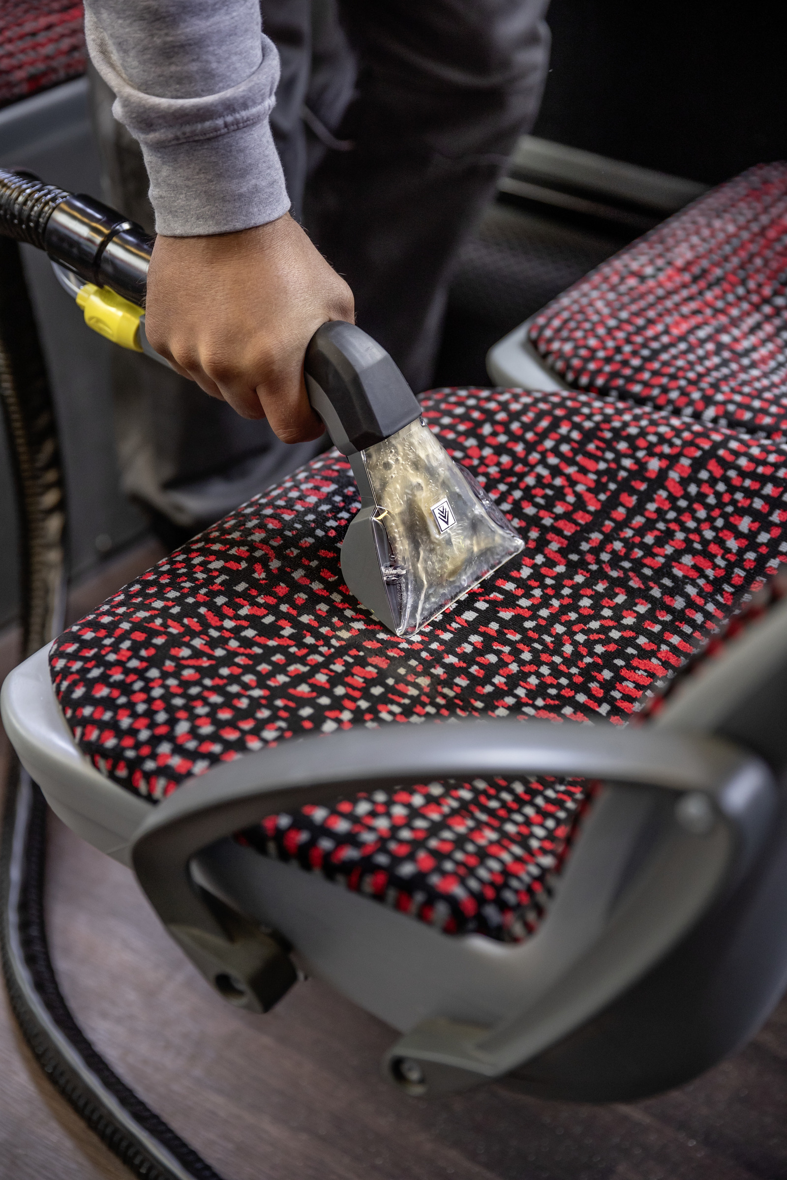 Osoba odkurza siedzenie autobusu czarno-żółtym odkurzaczem. Siedzenie ma czerwono-czarny wzór.