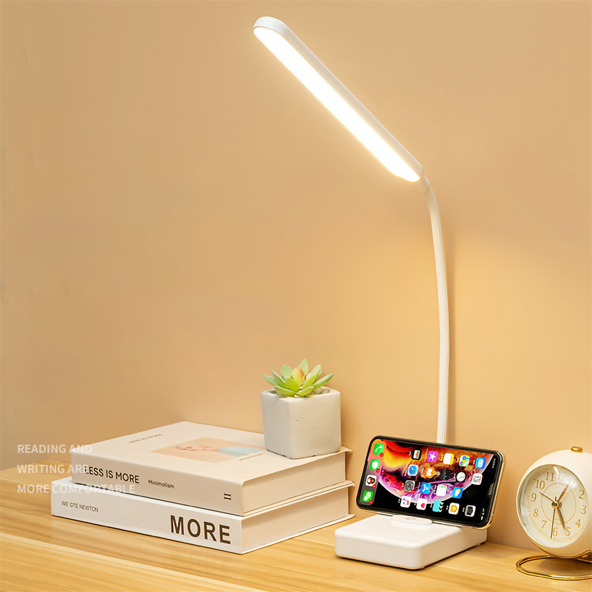 Eine weiße Schreibtischlampe mit Telefon-Ladeständer, einem Buch, einer Pflanze und einer Uhr.