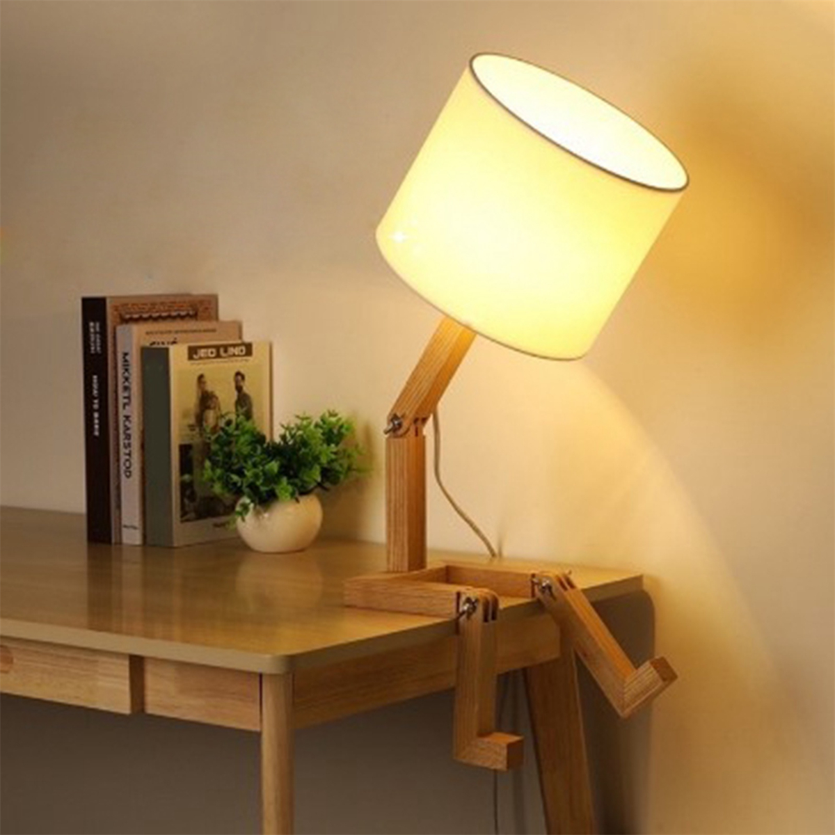 Eine holzfarbene Schreibtischlampe in Form einer Person mit weißem Lampenschirm, Büchern und einer Pflanze.