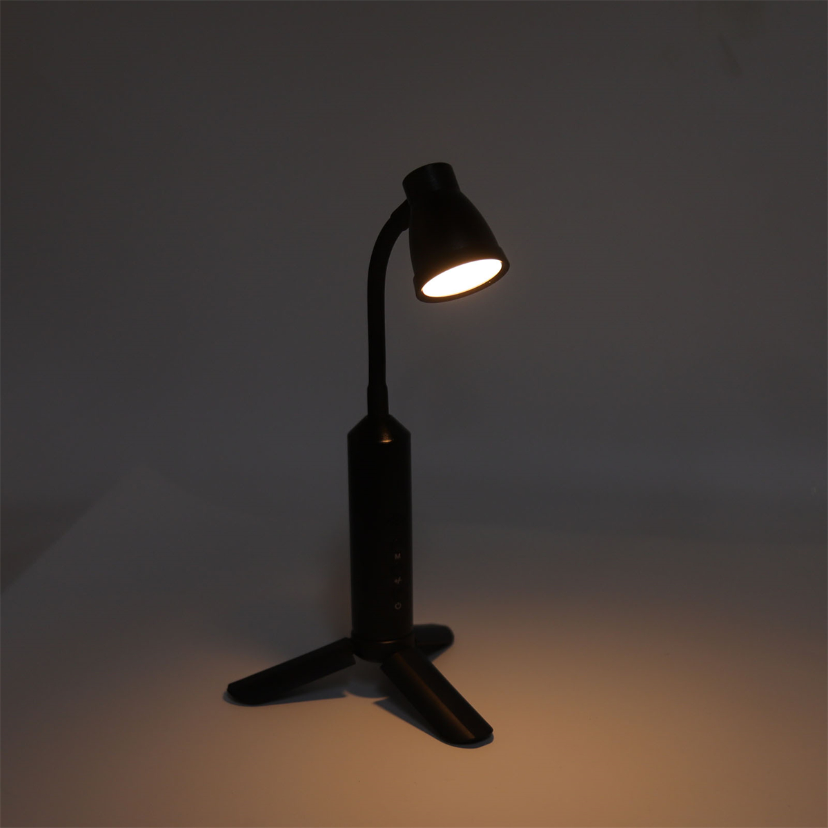 Schwarze Schreibtischlampe mit biegsamem Hals und eingeschaltetem Licht, vor dunklem Hintergrund.