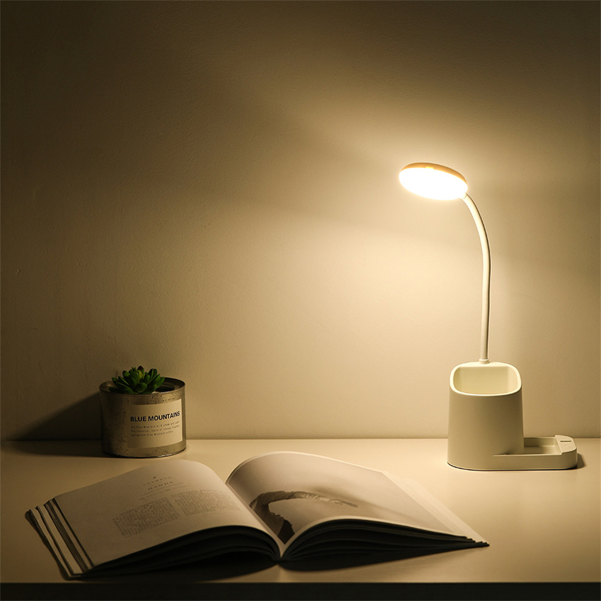 Eine Schreibtischlampe und ein offenes Buch auf einem Tisch. Es gibt eine kleine Pflanze.