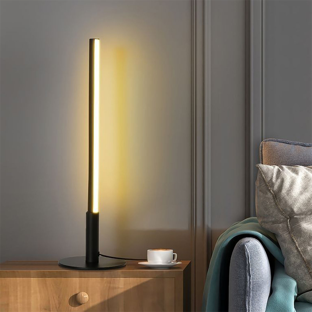 Eine hohe schwarze Lampe mit hellem vertikalem Licht auf einer Holzoberfläche. Eine Tasse Kaffee steht daneben.