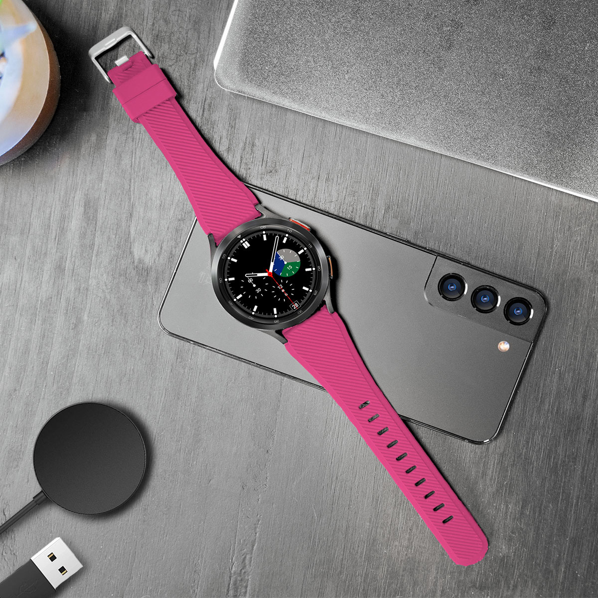 Eine Smartwatch mit pinkfarbenem Armband auf einem Telefon. Ein Laptop und eine Ladestation stehen auf dem Tisch.