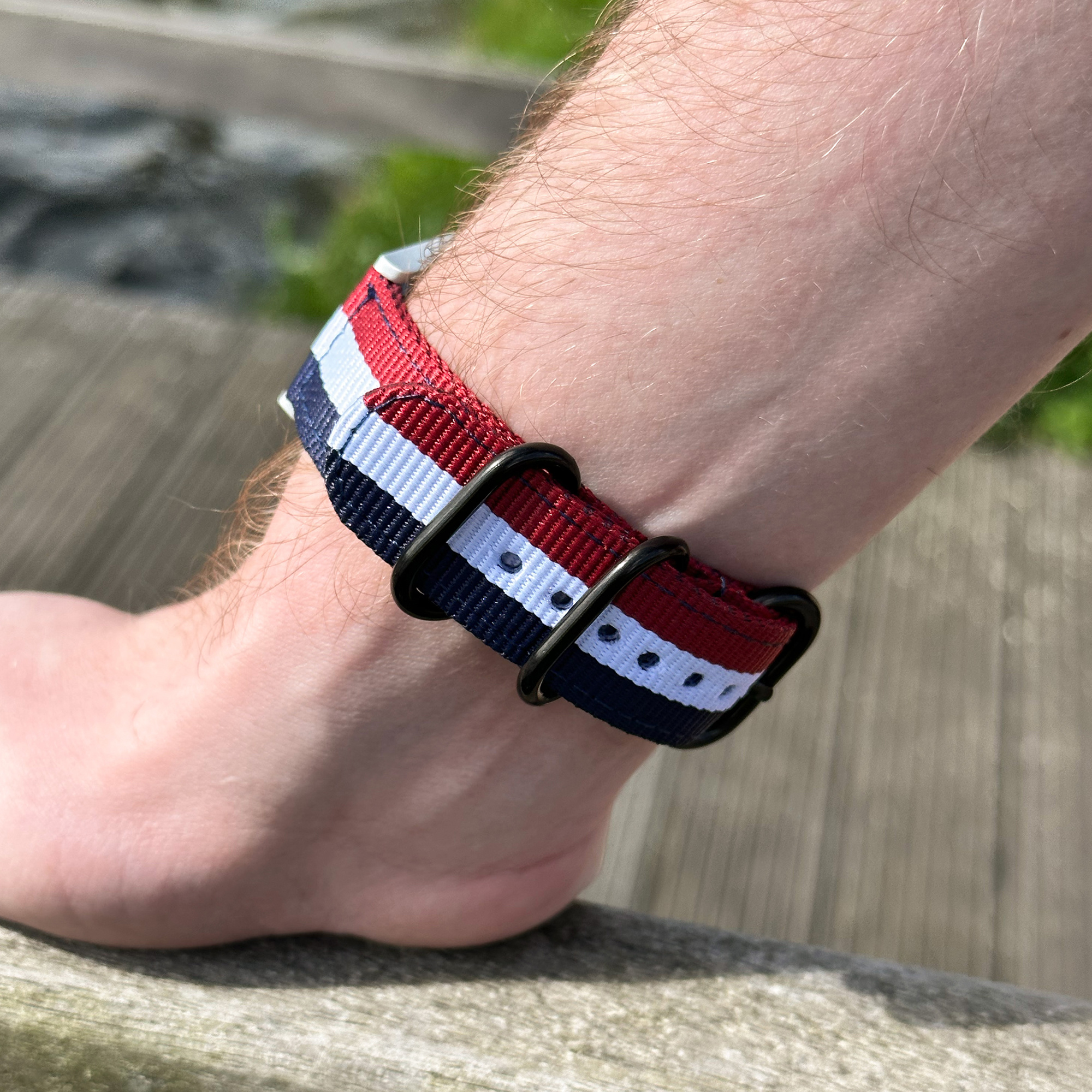 Handgelenk mit rotem, weißem und blauem Uhrenarmband. Schwarze Metallschlaufen. Holziger Hintergrund.