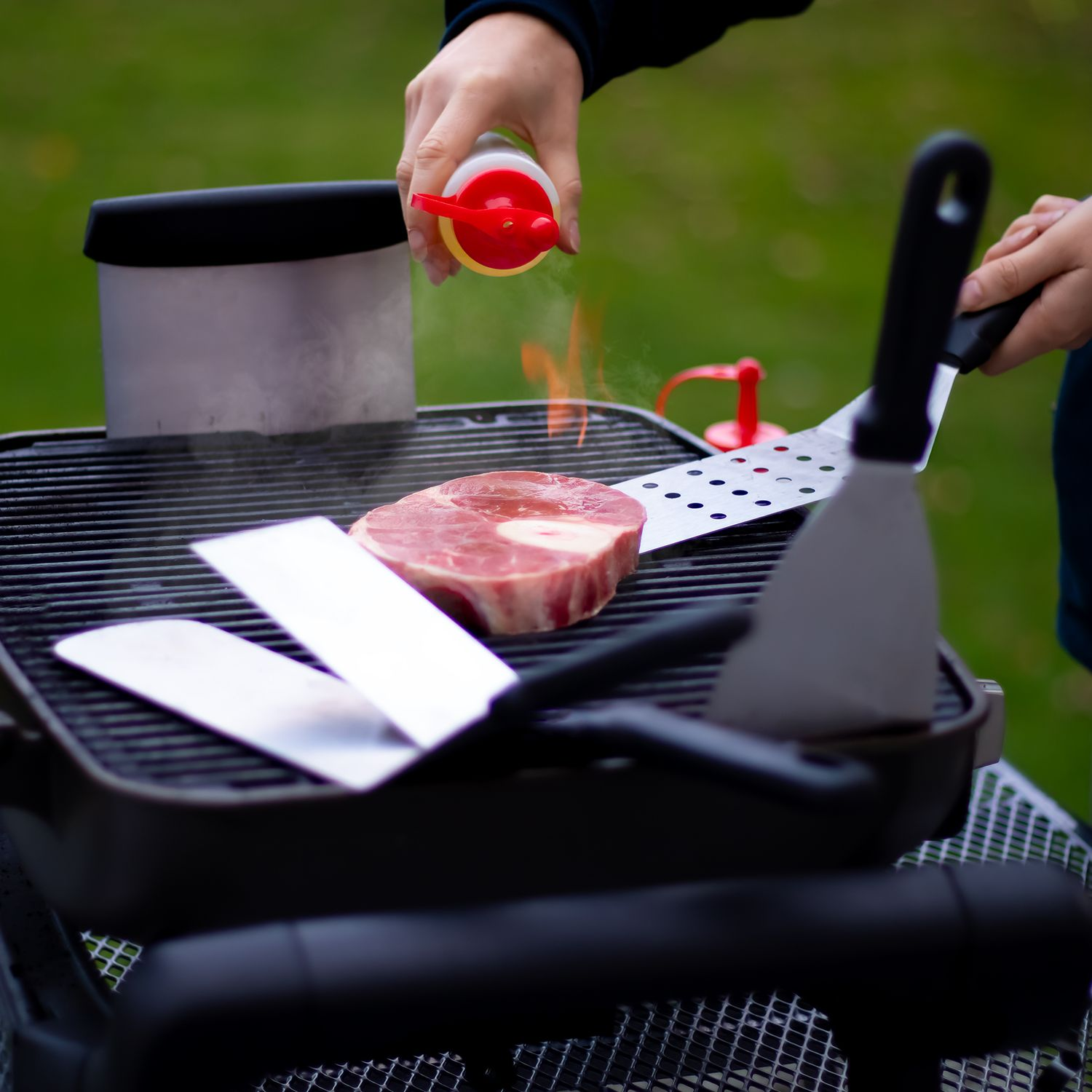 Ein Steak wird auf einem Grill mit einem Gewürz beträufelt. Spatel und ein Behälter sind in der Nähe.