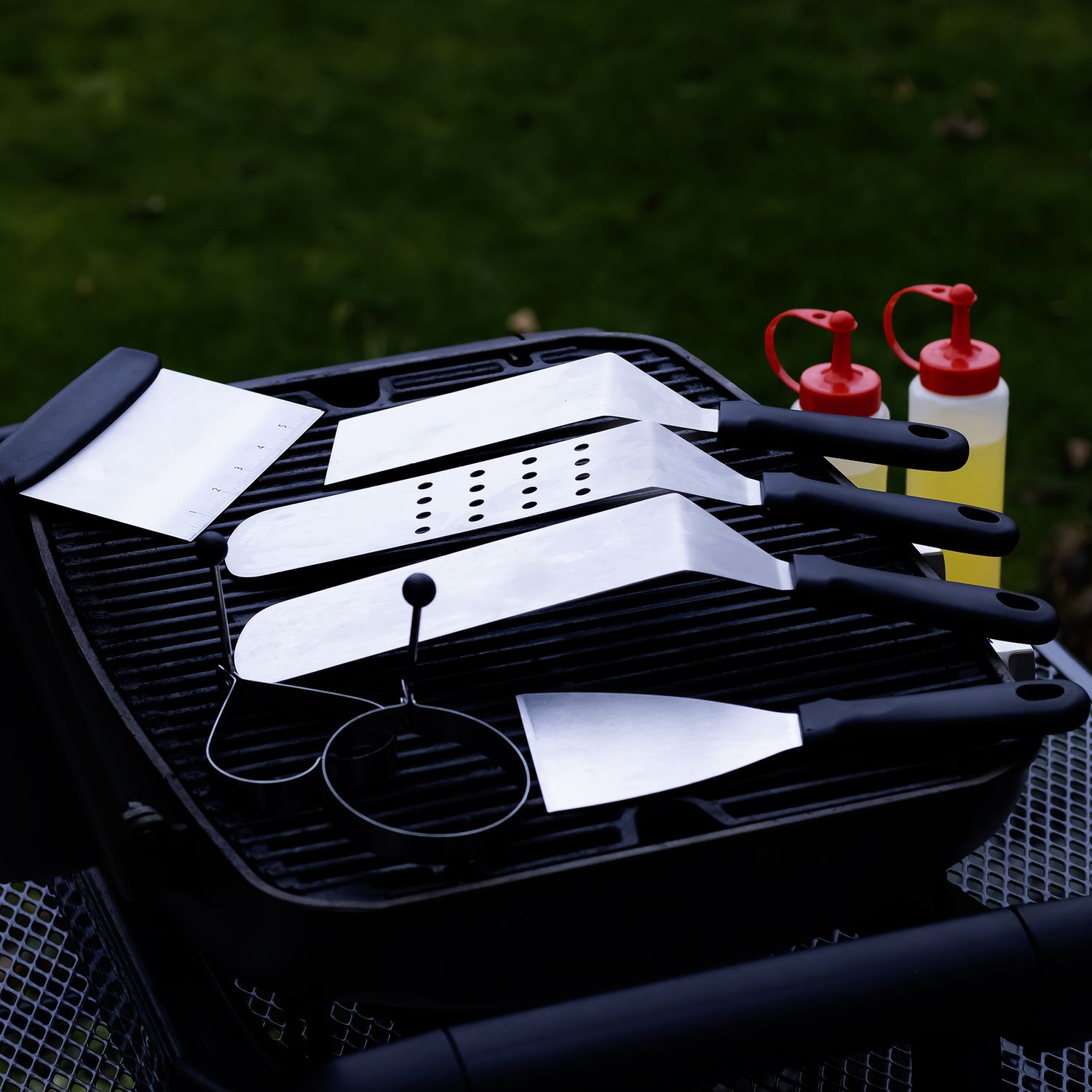 Grill mit Grillwerkzeugen, einschließlich Spateln und Zangen. Rote und gelbe Flaschen sind sichtbar.