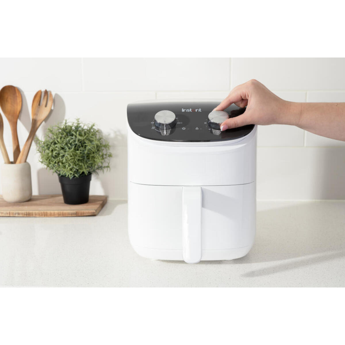 Een witte airfryer met een hand die aan een knop draait. Keukengerei, houten lepels en een plant links. Wit aanrecht.