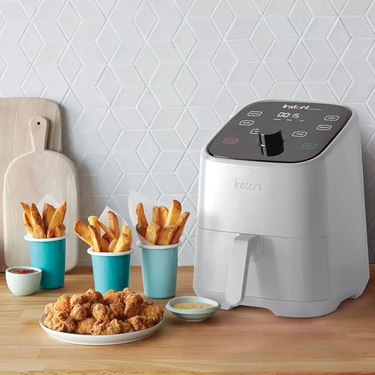 Friggitrice ad aria con patatine fritte e pepite di pollo su un tavolo. Un tagliere è visibile sullo sfondo.