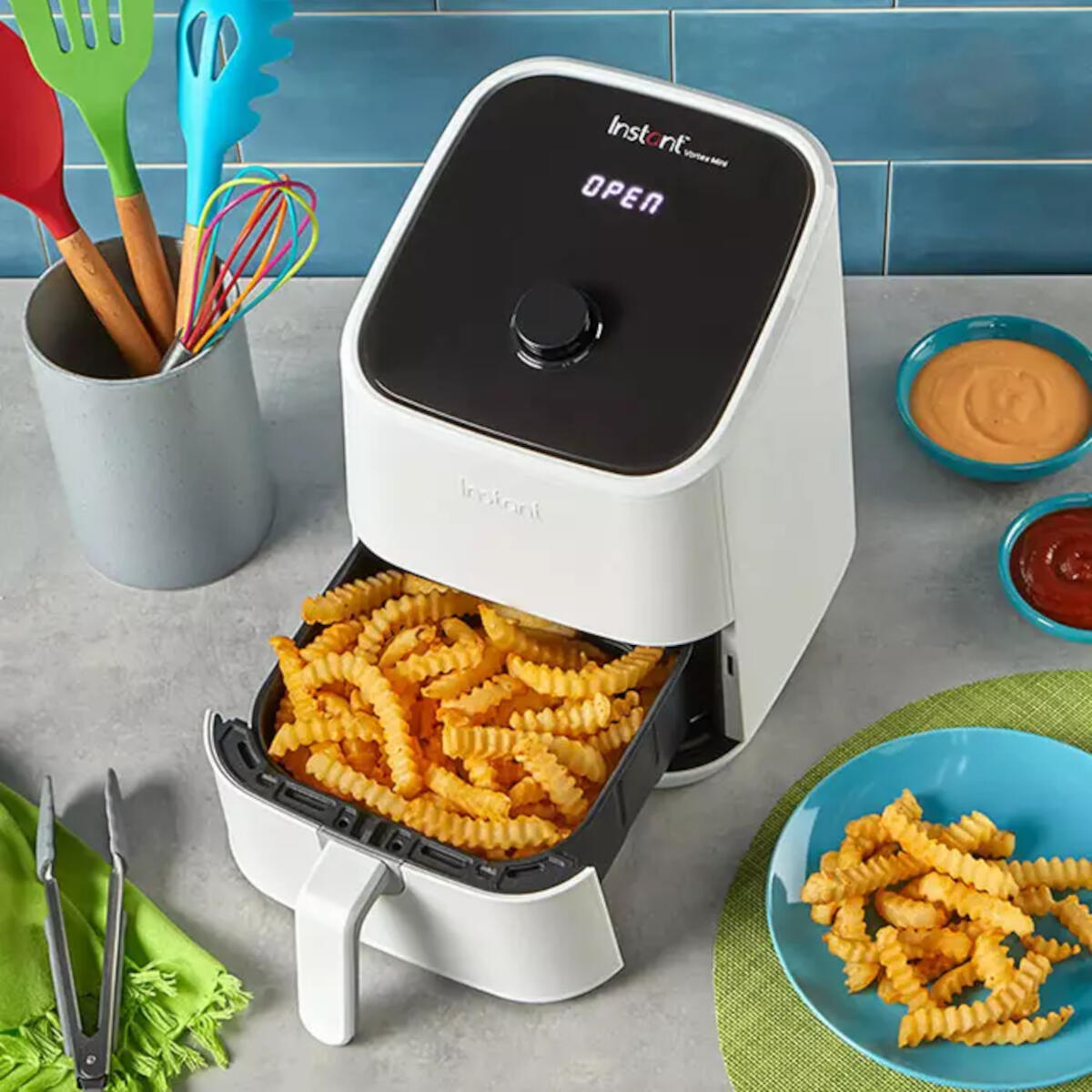 Friggitrice ad aria bianca con patatine fritte, salse e utensili su un tavolo blu.