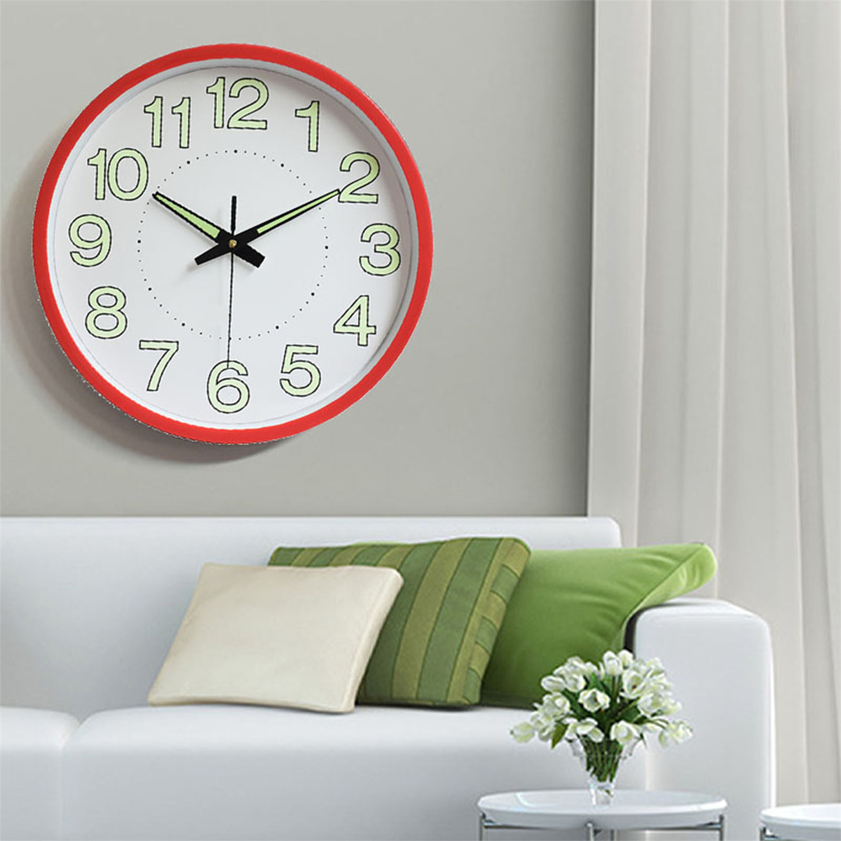 Eine weiße Uhr mit rotem Rand, schwarzen Zeigern und grün leuchtenden Ziffern. Die Uhr hängt an einer grauen Wand.