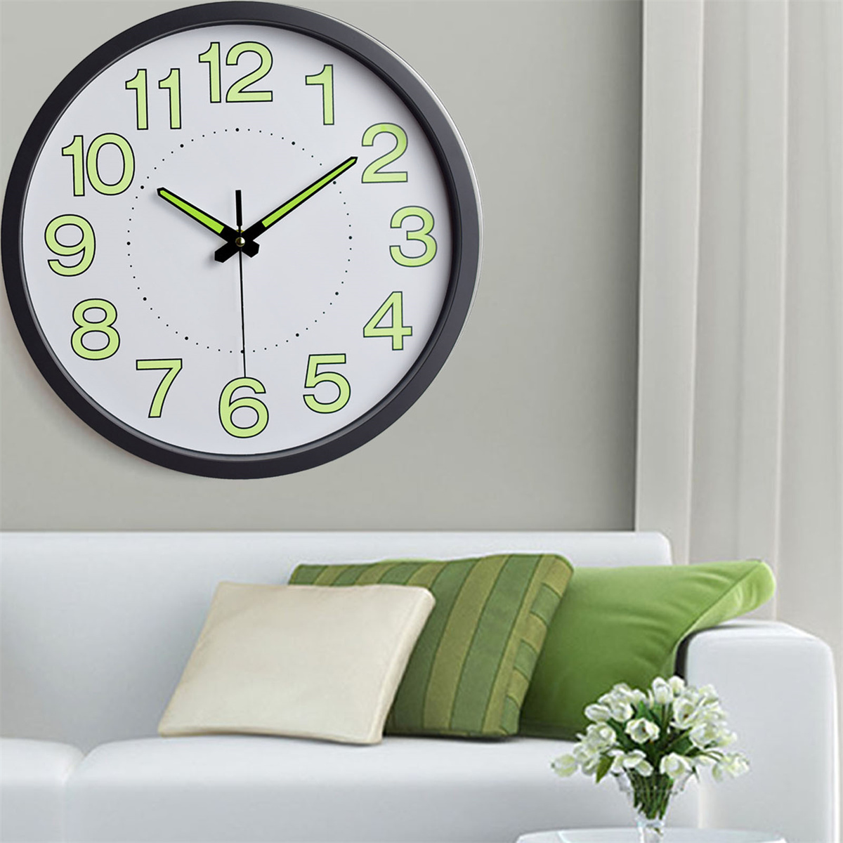 Eine Uhr mit schwarzem Rahmen. Sie hat ein weißes Zifferblatt mit grünen Zahlen und Zeigern und befindet sich über einem weißen Sofa.