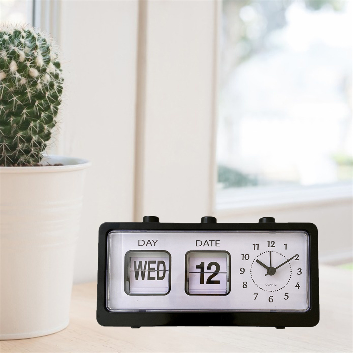 Schwarze Retro-Uhr mit Tages- und Datumsanzeige. Im Hintergrund ein Kaktus in einem weißen Topf.