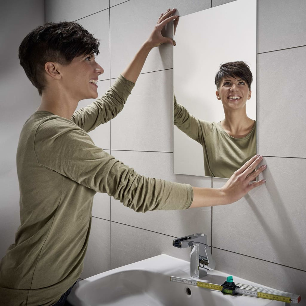 Frau hängt Spiegel an der Wand, lächelt. Eine Wasserwaage befindet sich unter dem Waschbecken.