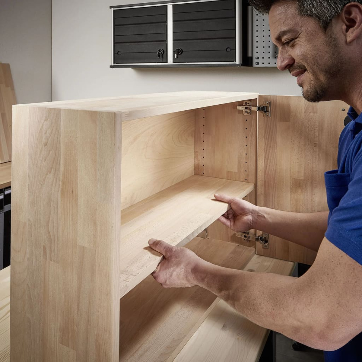 Mann montiert Holzschrank, hält ein Regal im Inneren. Der Schrank steht in einer Werkstatt.