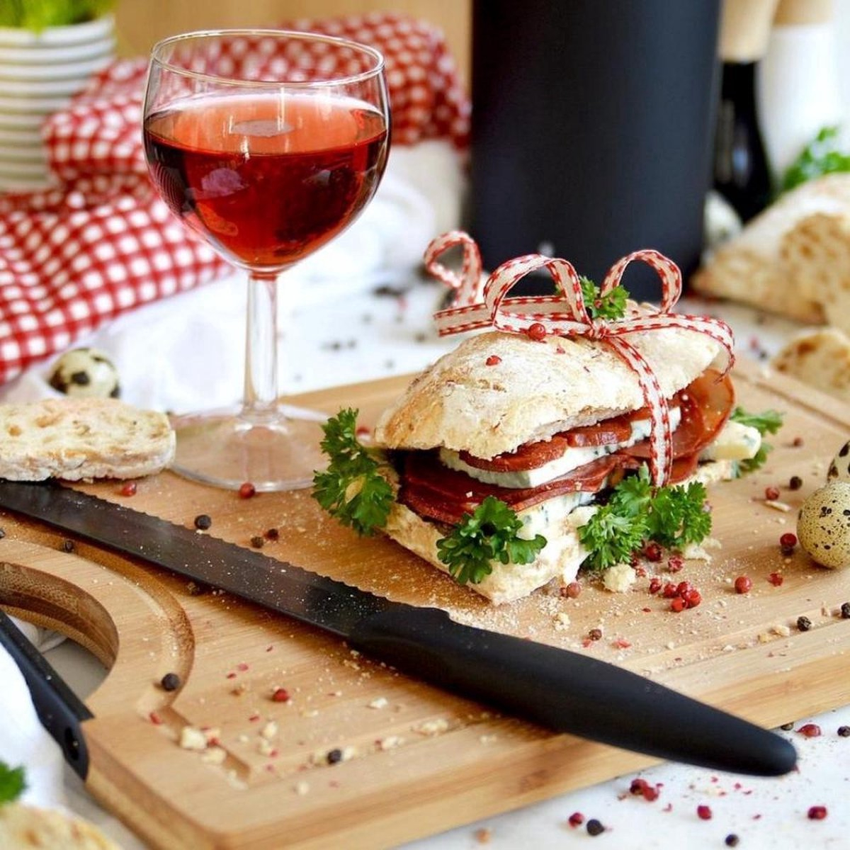 Kanapka z mięsem i serem, kieliszek czerwonego wina i nóż na drewnianej desce z dodatkami.