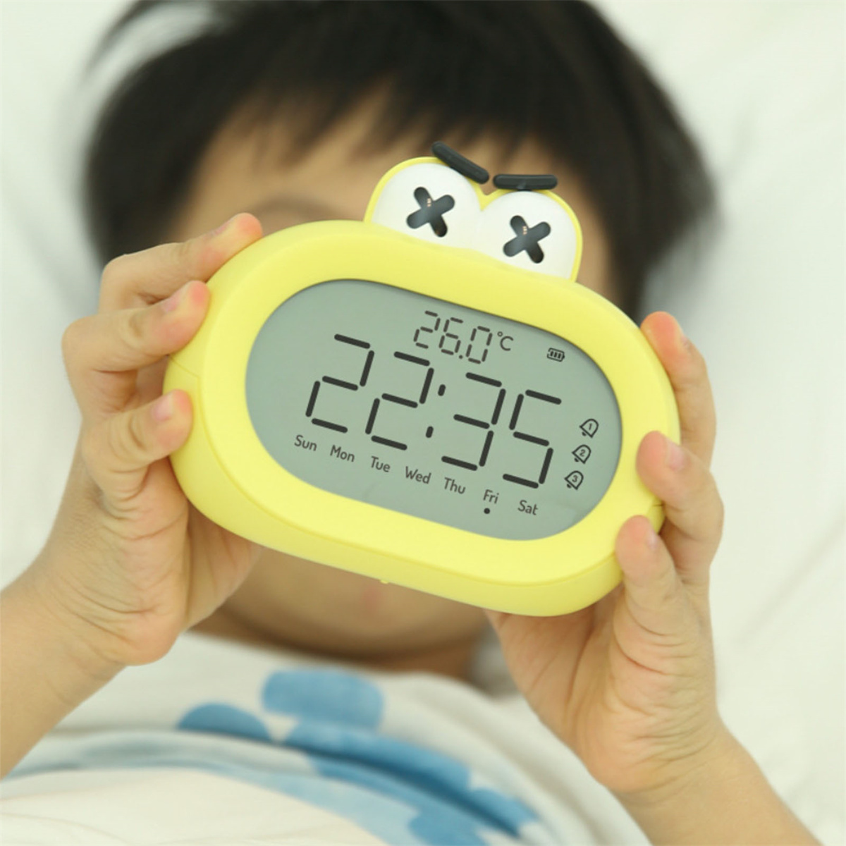 Ein Kind hält einen gelben Wecker. Die Uhr zeigt 22:35 und 26,0 Grad Celsius an.