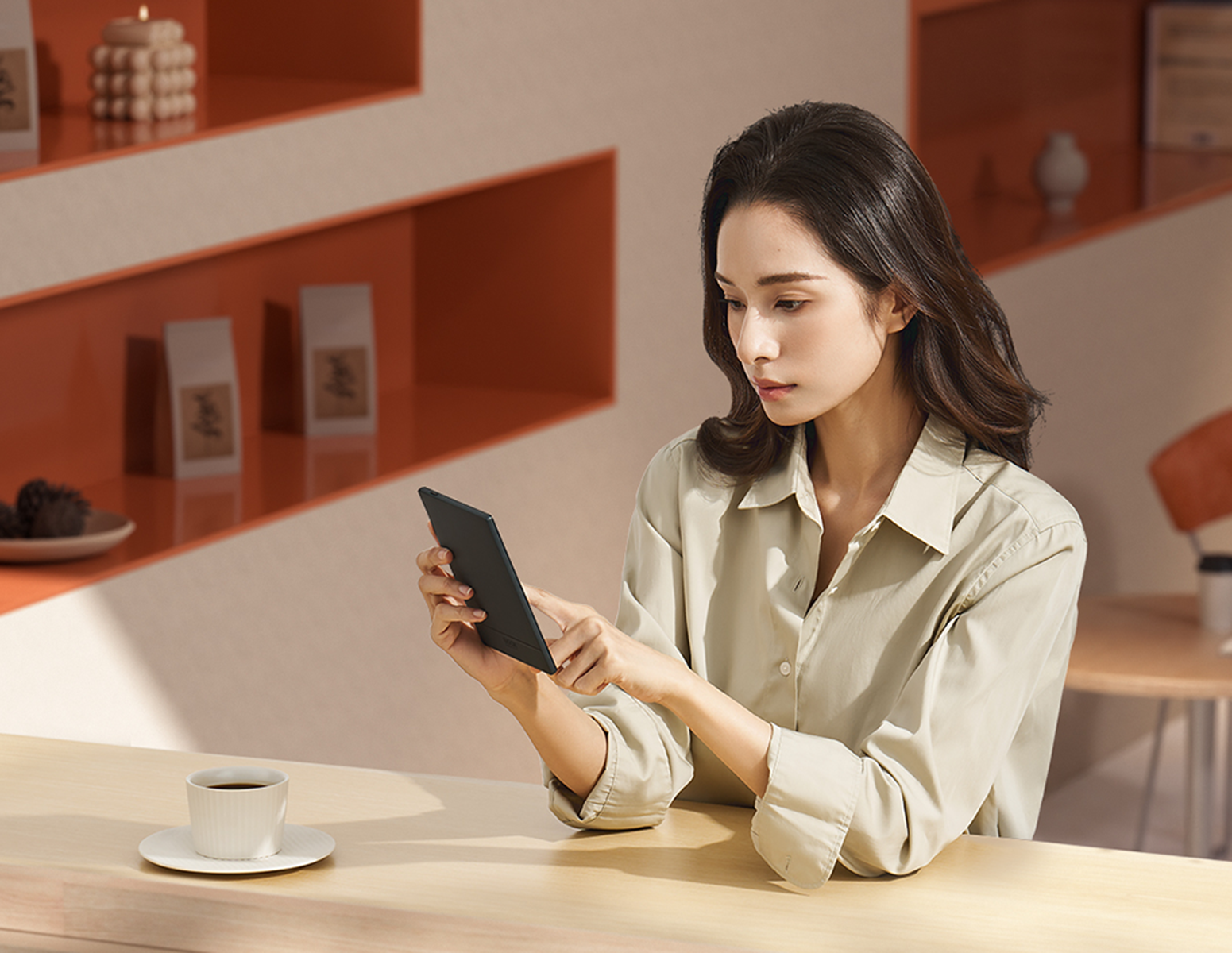Kobieta używa czytnika e-booków, trzymając go w rękach. Siedzi przed filiżanką kawy na stole.