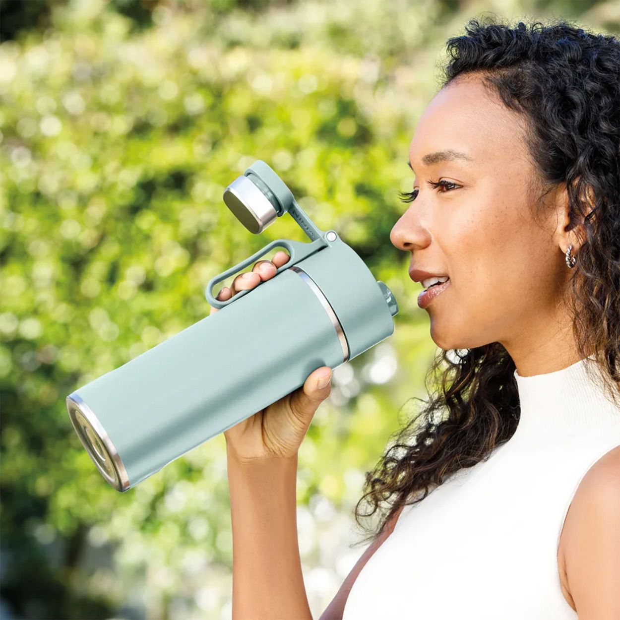 Una donna beve da una borraccia verde acqua con un manico. È in un ambiente esterno.
