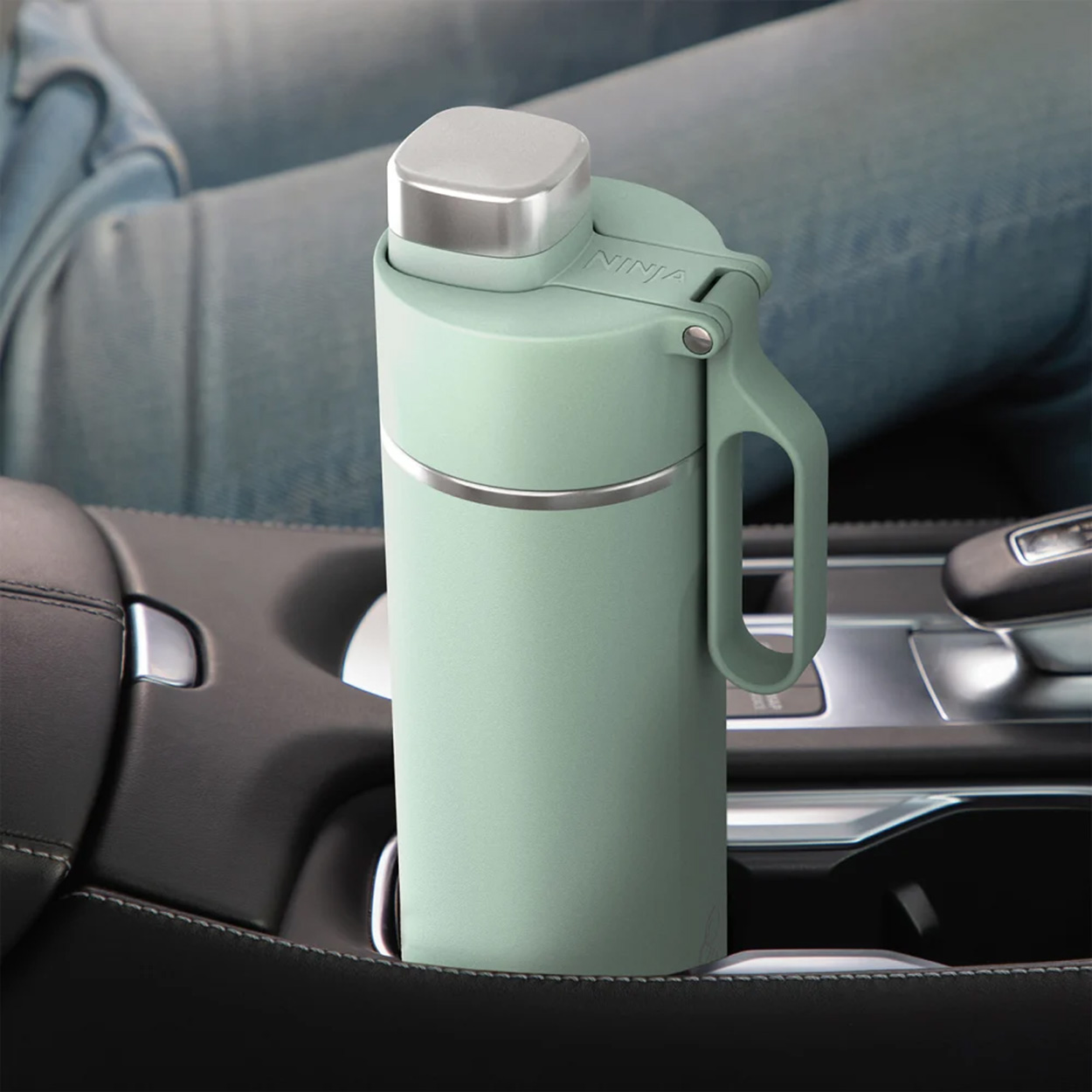Tazza da viaggio verde con manico, in un portabicchieri per auto. Jeans sullo sfondo.