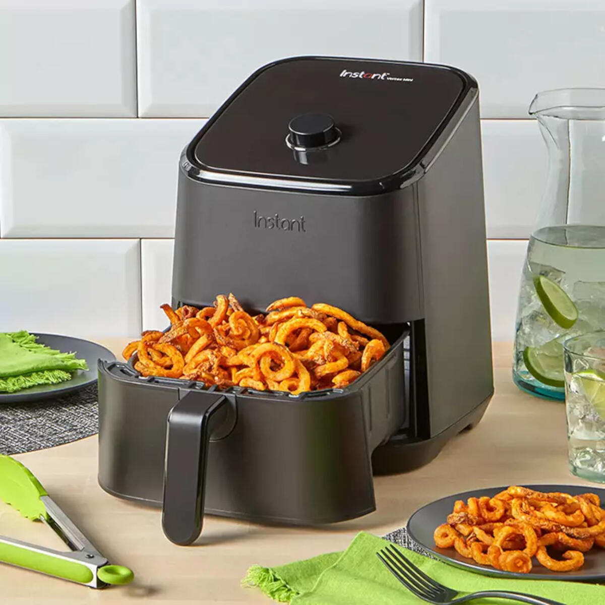 Zwarte airfryer met curly fries in de mand. Andere borden met friet en een kan op een tafel.
