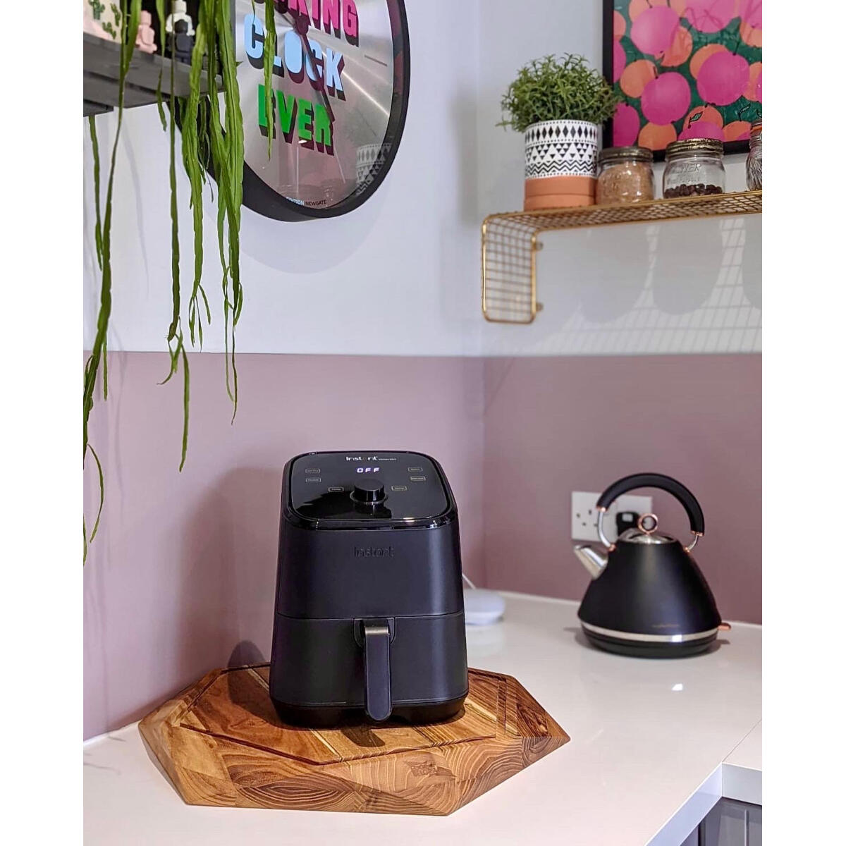 Zwarte airfryer op houten plank. Keuken met wit aanrecht, roze muur en waterkoker.
