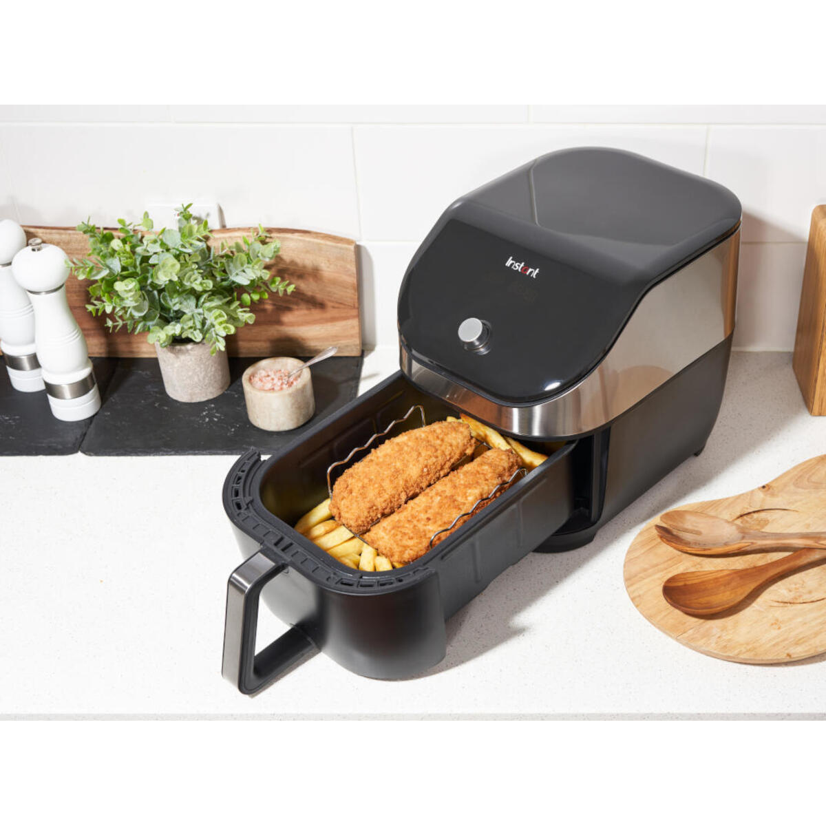 Een airfryer met eten erin, op een aanrecht met keukengerei en planten in de buurt.