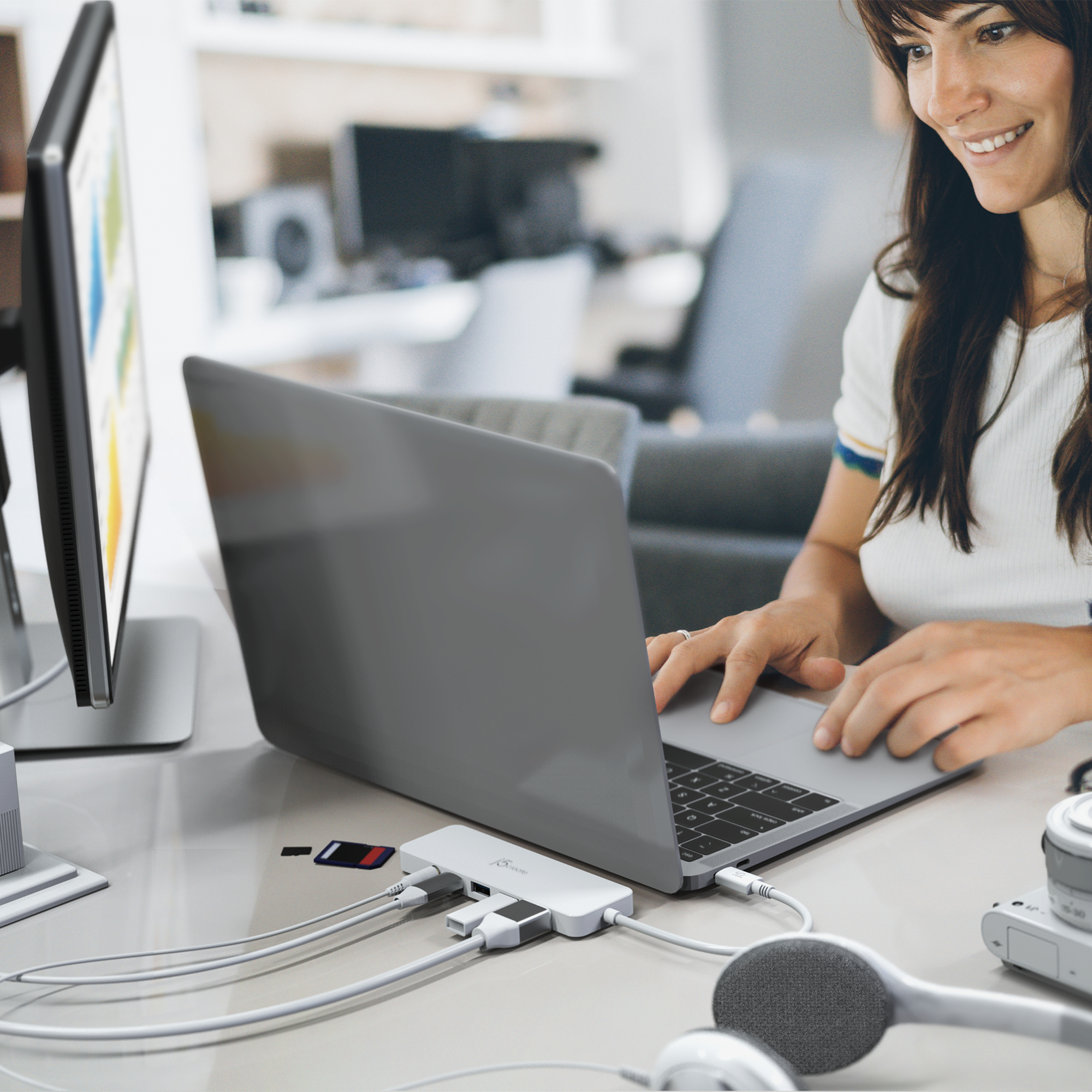 Eine Frau benutzt einen Laptop, der mit einem Hub verbunden ist. In der Nähe befinden sich Kopfhörer, ein Monitor und eine Speicherkarte.