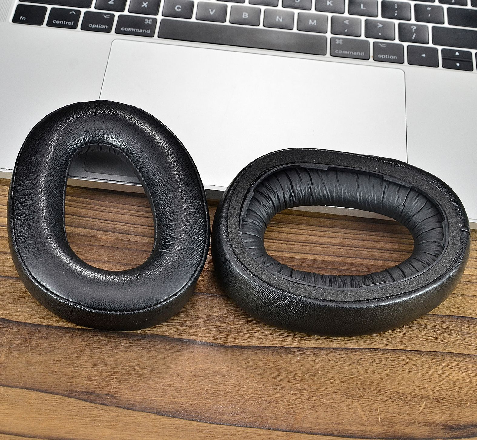 Twee zwarte oorkussens voor een hoofdtelefoon op een houten oppervlak met een laptop op de achtergrond.